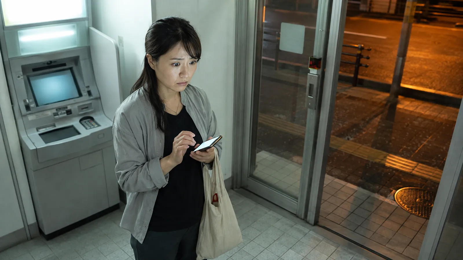 夕方のATMコーナーで、請求額より少ない入金をスマートフォンで見つけて立ち尽くすフリーランス